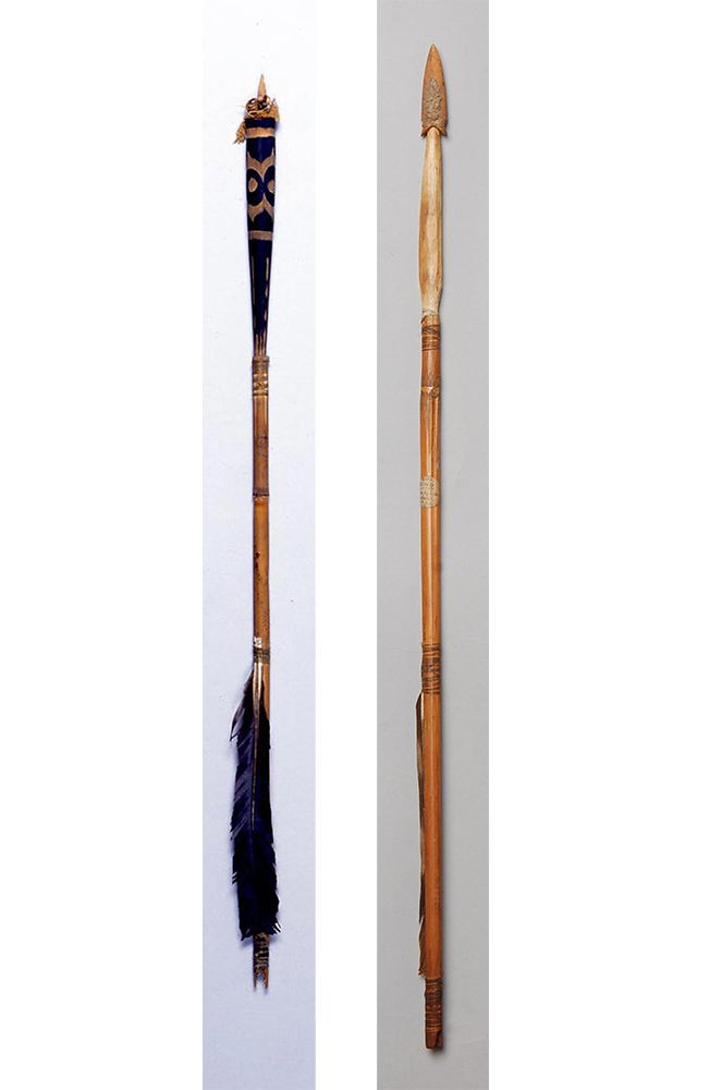 Two arrows next to each other vertically. On the left is a blunt-tipped arrow with dark patterns on the tip and blackish-blue feathers. On the right is a lighter wooden arrow with thinner feathers and a pointed tip.