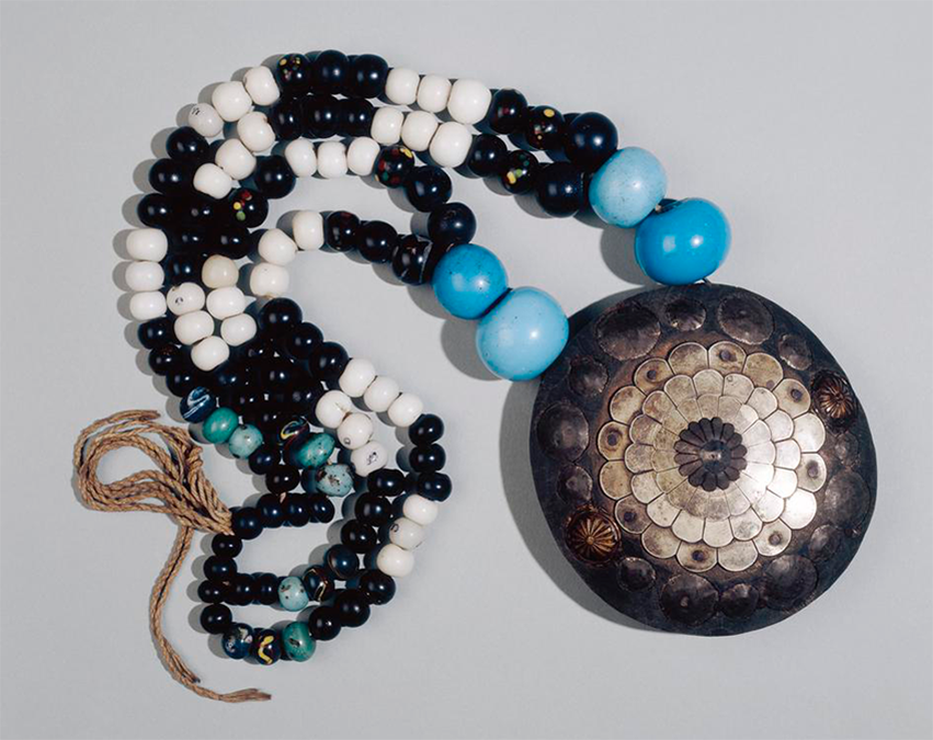 A traditional tamasay necklace with beads of black, white, and blue with two larger blue beads on either side of a giant silver medallion with circular patterns on it.
