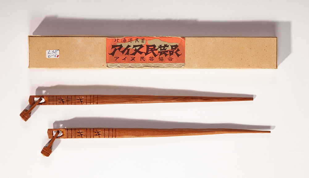 A long horizontal cardboard box with a red label and Japanese writing. Below it is a pair of carved chopsticks with a little carved bell shape on the end.