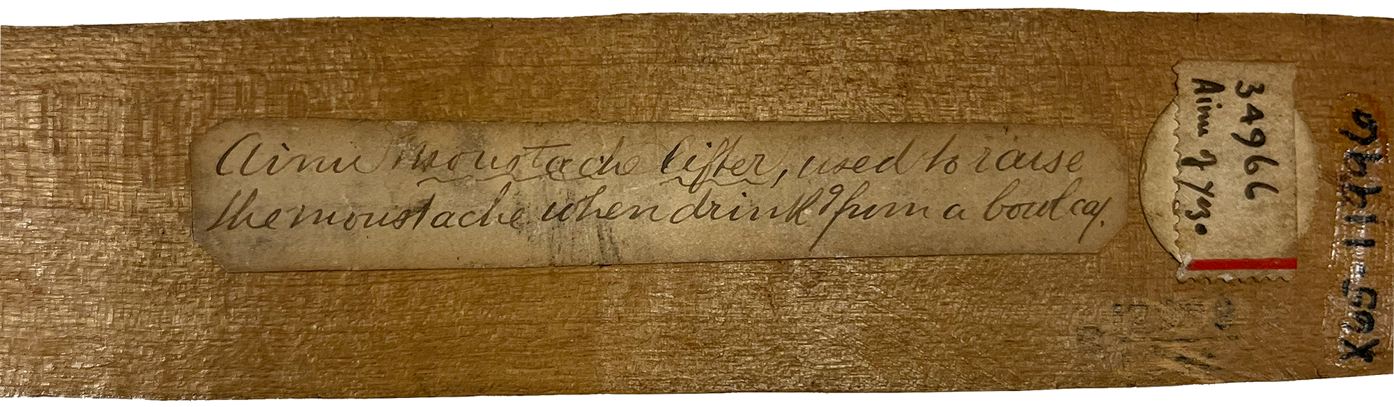 Closeup of the back of a carved wooden stick with an aged label in cursive that says 'Ainu moustache lifter, used to raise the moustache when drinking from a bowl.'