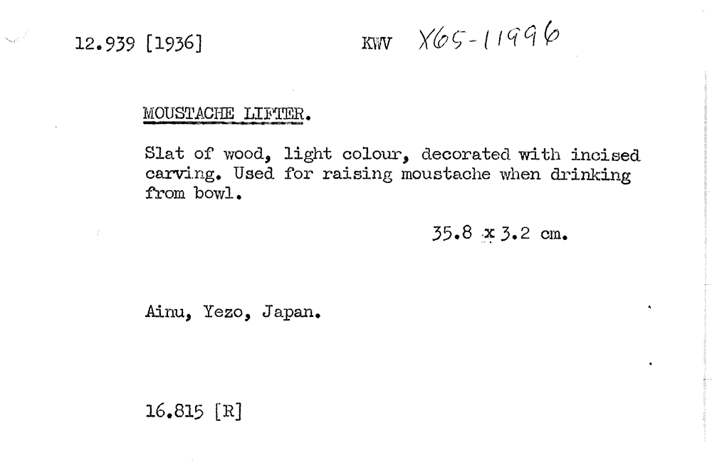 A provenance record of typed text that says 12.939 [1936] KWV X65-11996 MOUSTACHE LIFTER. Slat of wood, light colour, decorated with incised carving. Used for raising moustache when drinking from bowl. 35.8 x 3.2 cm. Ainu, Yezo, Japan. 16.815 [R]