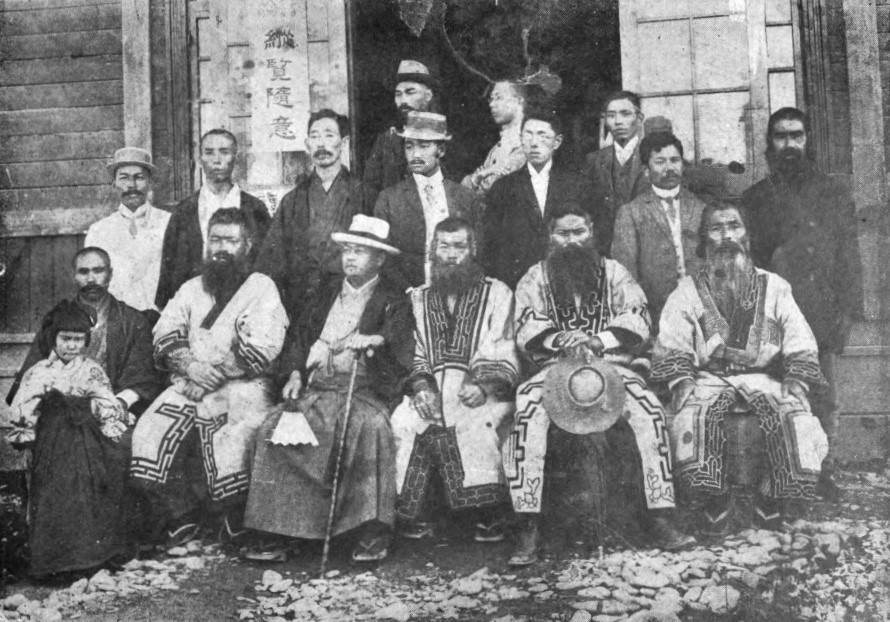 a black and white photograph of over a dozen Japanese and Ainu men, some dressed in suits and hats in a modern style and the Ainu men in traditional Ainu patterned robes.