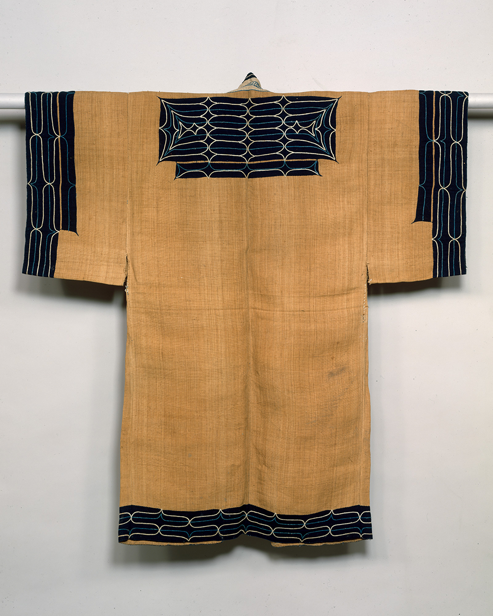 A photo of an Ainu attush robe with a light brown main color and dark bluish black patterns on the edges of the sleeve and hems that have white geometric designs.