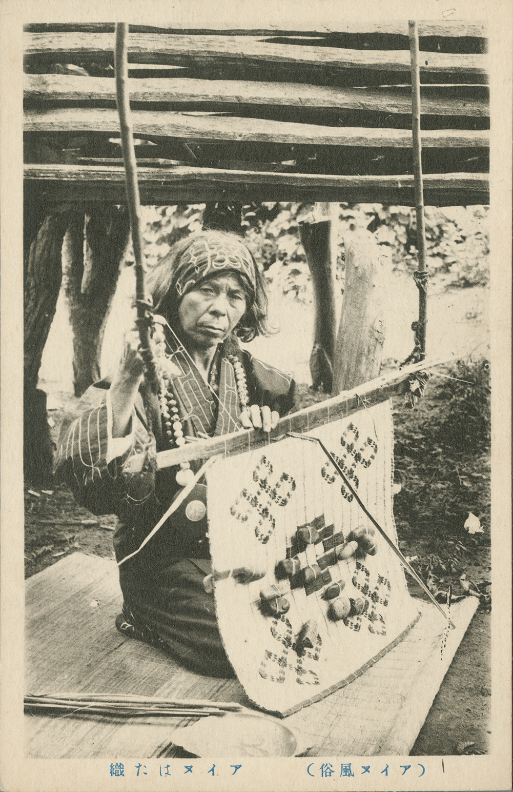 A postcard photograph of an older Ainu woman kneeling on a mat as she uses an itaseni loom to weave.