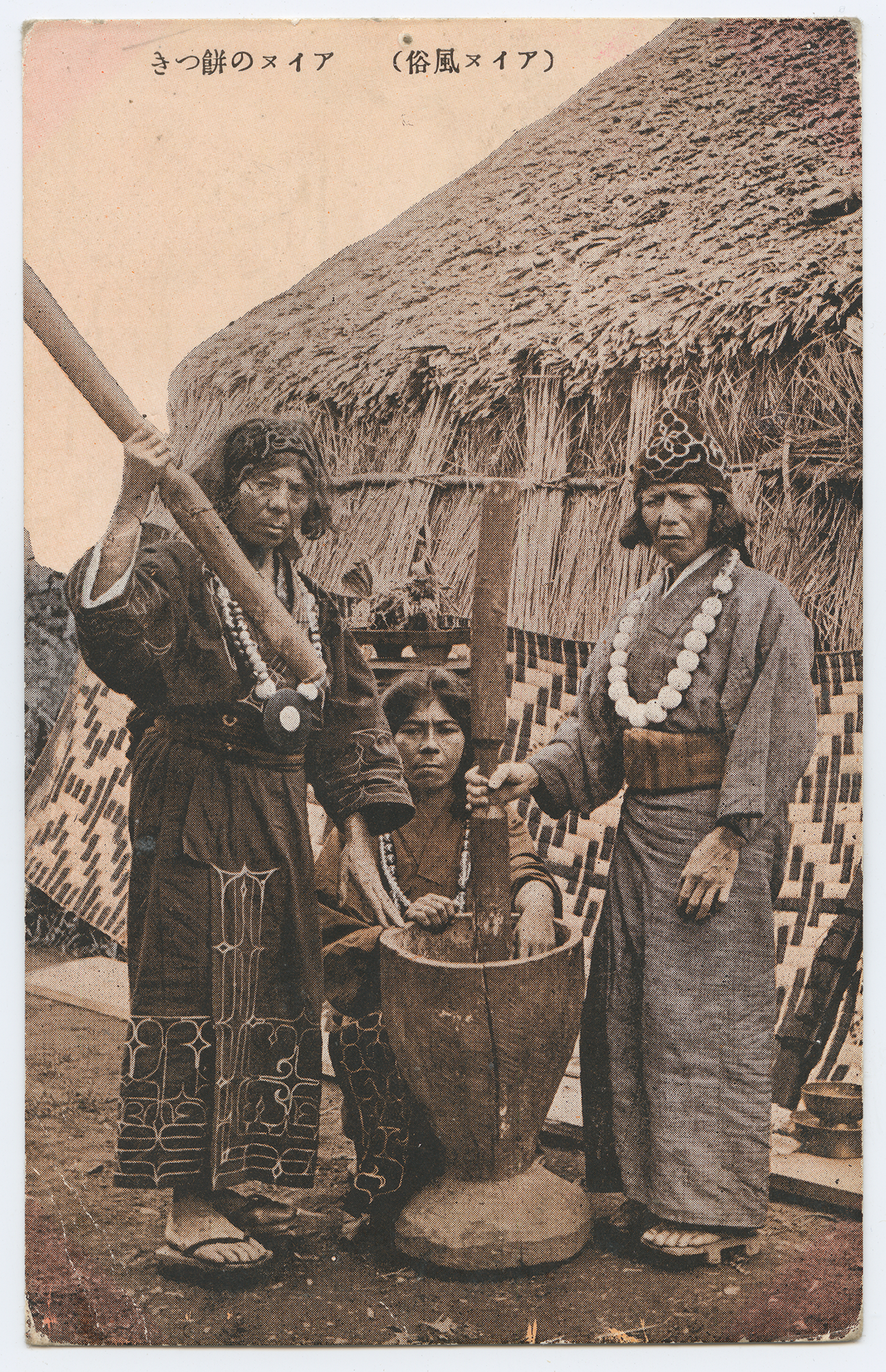 A photograph on a postcard in sepia tones of three Ainu women in front of a large wooden mortar, two of them holding pestles. They are in traditional Ainu dress and staring at the camera.