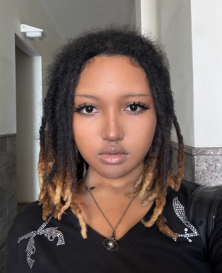 A Black Asian woman in makeup with a nose ring. Her dark hair has light tips and she is wearing a black shirt with sparkly designs. Her expression is neutral.