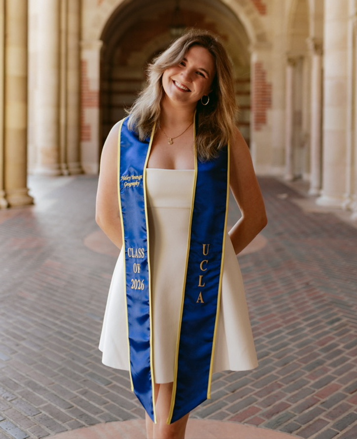 A white woman with blonde hair smiling and standing in an outside hall. She has on a white dress and a blue sash that says UCLA